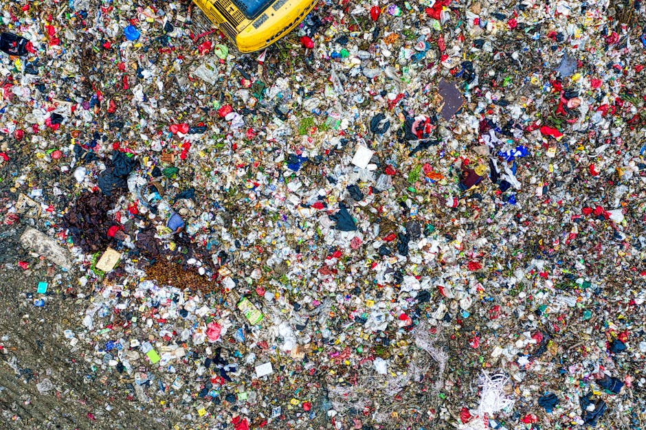 Aerial view of a large, densely packed landfill site filled with a mix of municipal waste including plastic bags, food wrappers, cardboard, textiles, and miscellaneous debris. The waste is scattered a