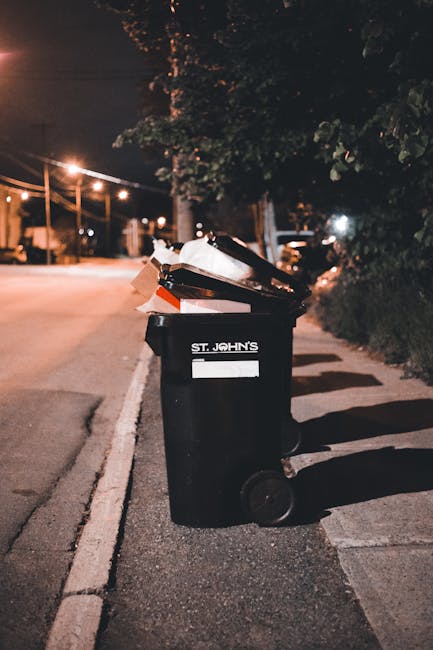 A row of five large wheeled rubbish bins made of durable black plastic with yellow lids, positioned against a plain light gray wall on a concrete surface. Each bin features a white oval label with ide